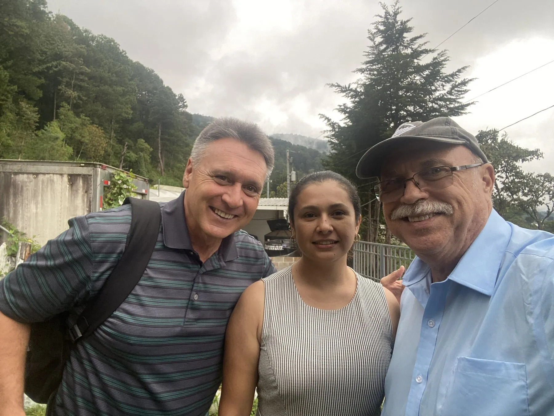 Paul standing with a Honduran pastor and his wife outdoors among pine trees.
