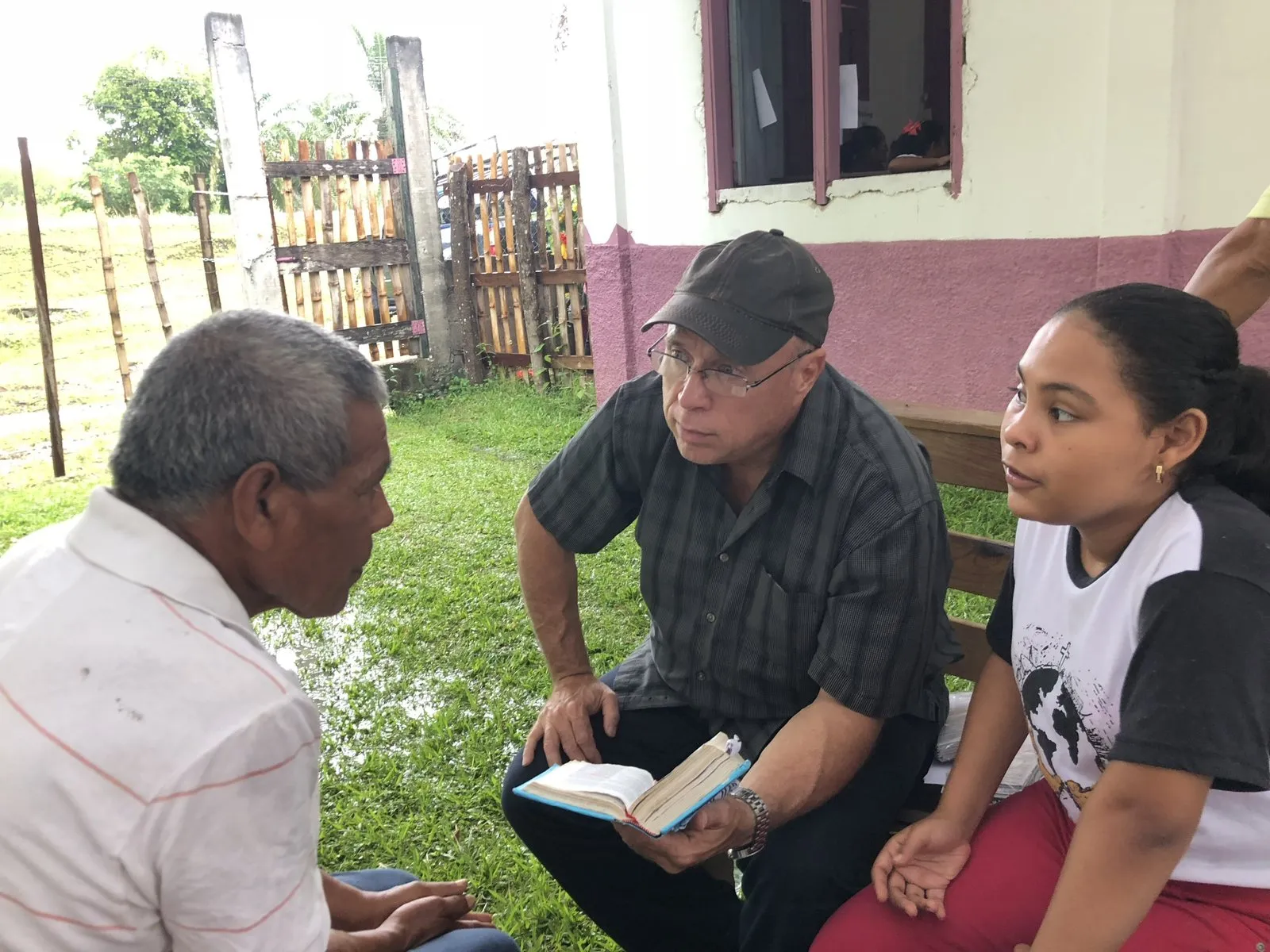 Paul seated outside a Honduran home with his Bible, talking with a man and a young woman.
